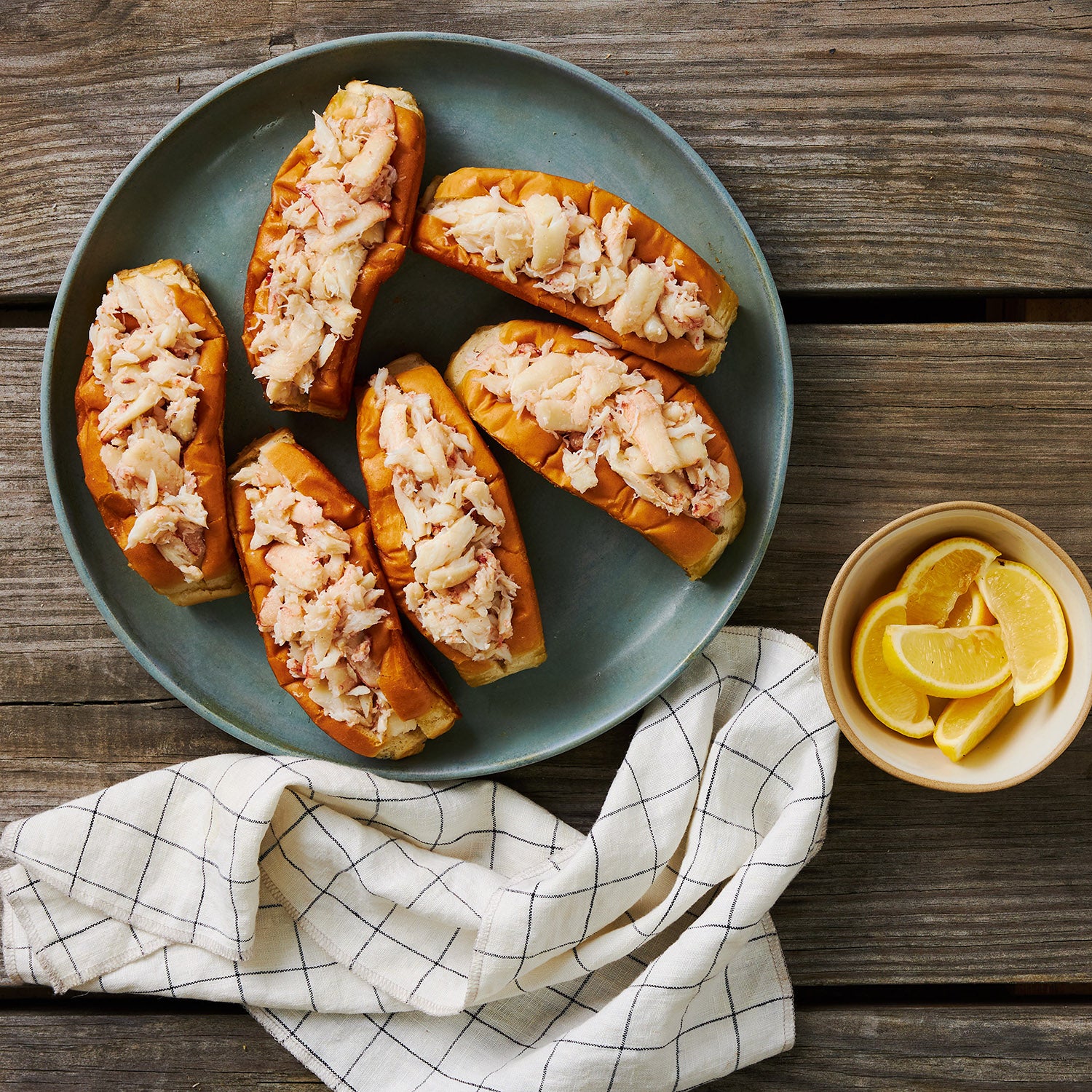 This Photograph shows the Six completed crab rolls out of the At-Home Kit plated beautifully beside a bowl of Lemon wedges, on top of a beautiful Wooden plank table.  The crab is teeming out of the Split top New england style rolls that are included in this kit to ensure you can have that authentic New England feel no matter where you choose to enjoy our At-Home Crab Roll Kit