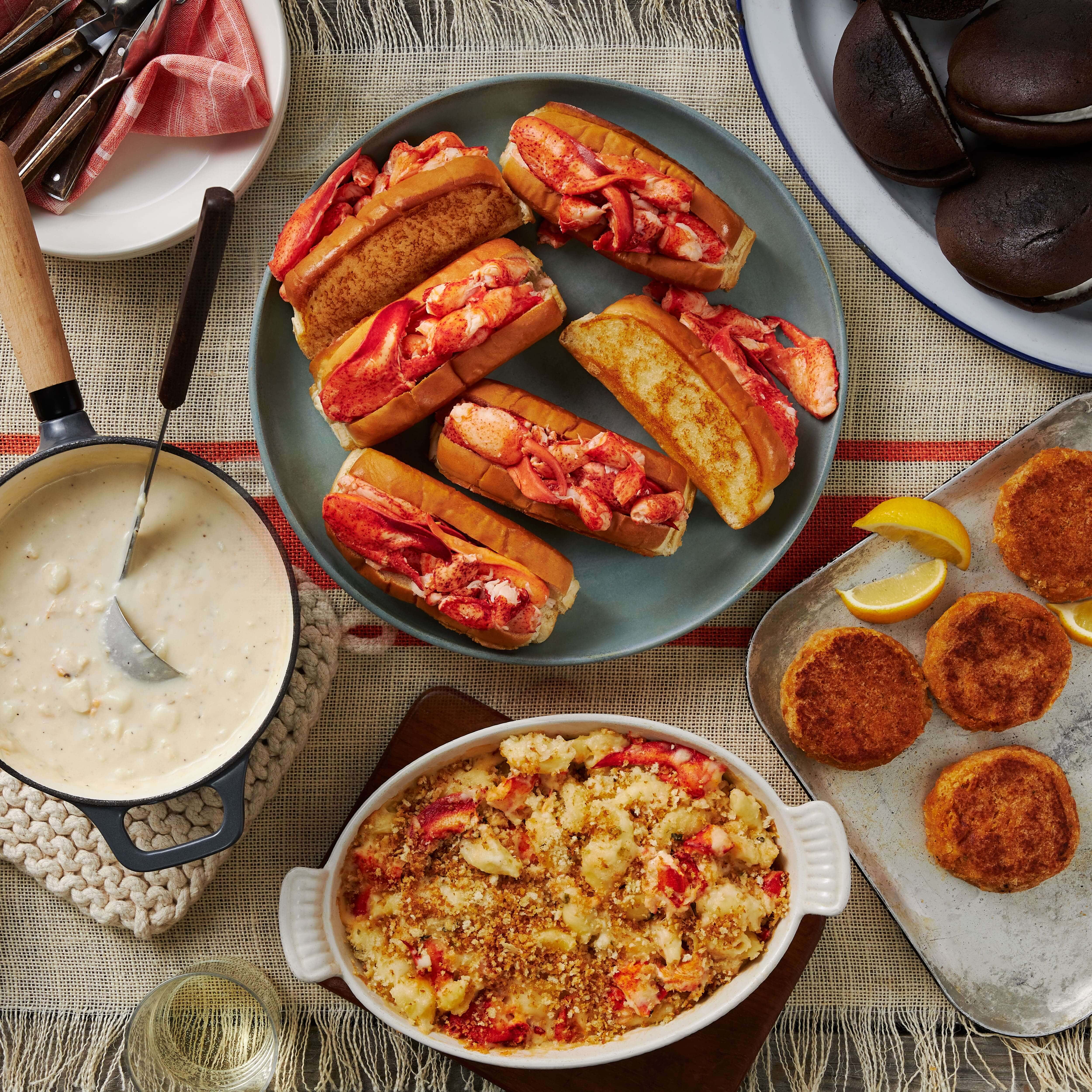 A variety of prepared seafood dishes including Maine lobster rolls, crab cakes, and whoopie pies, along with bowls of clam chowder and lobster bisque.