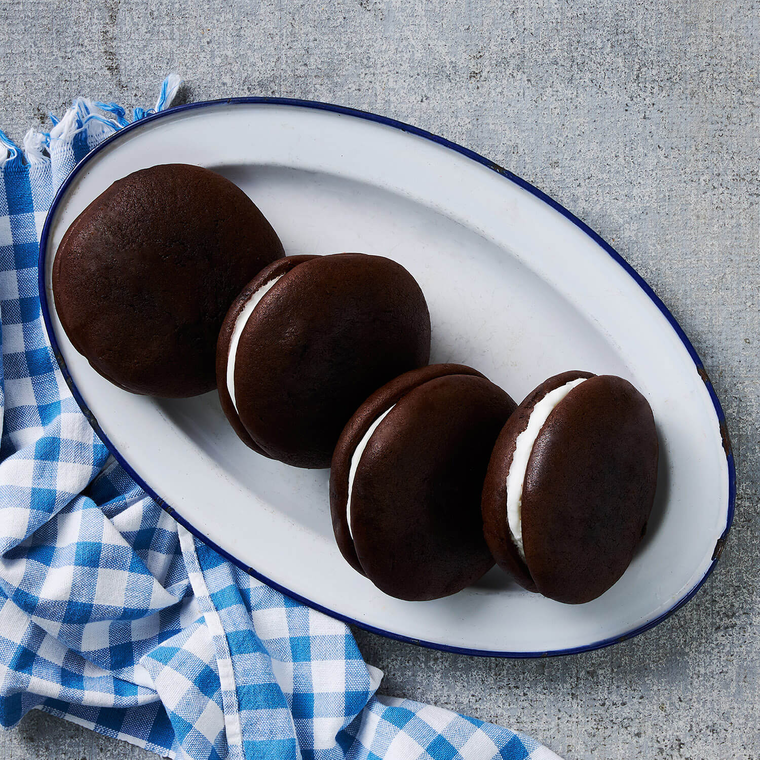 A plate with four chocolate whoopie pies, two with filling facing up, served on a white plate with a blue rim on a concrete surface, accompanied by a blue and white checkered napkin.
