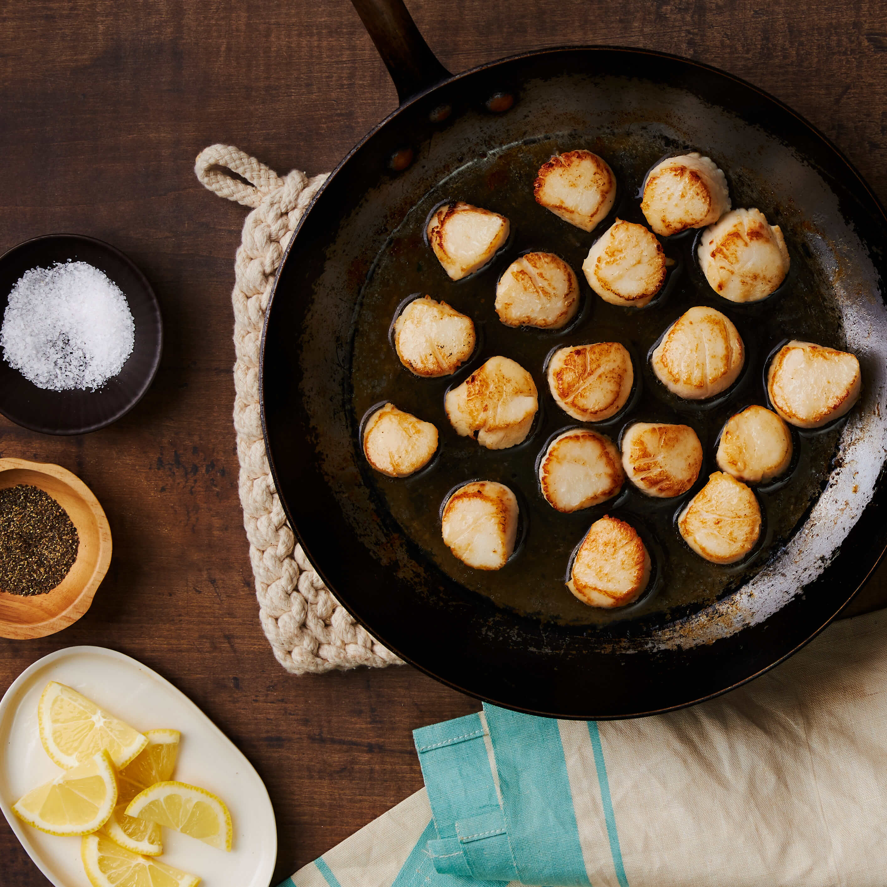 Cooked Scallops in a frying pan on a wooden table with lemon slices and salt.