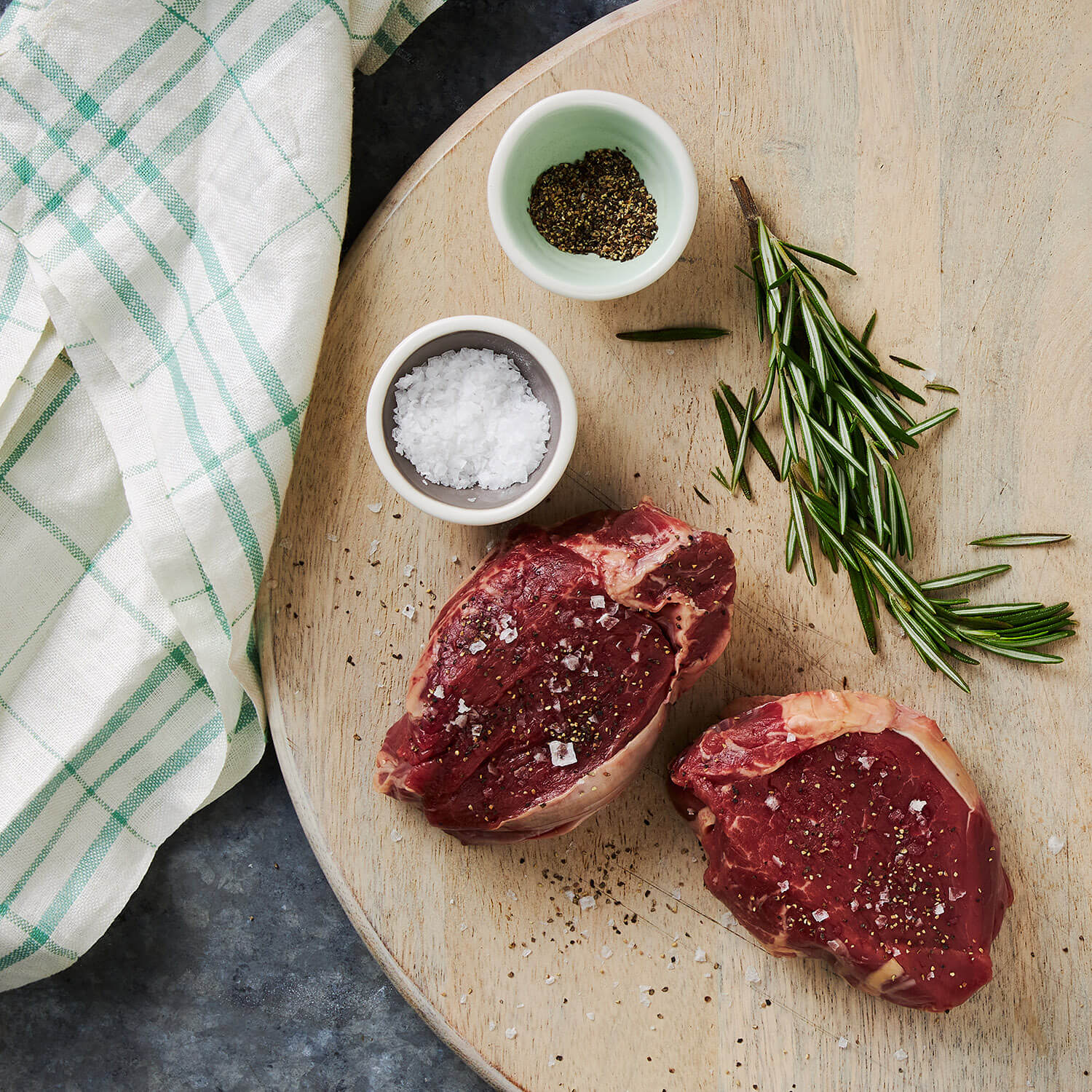 Two raw tenderloin steak fillets on a wooden cutting board with salt, pepper, and rosemary.