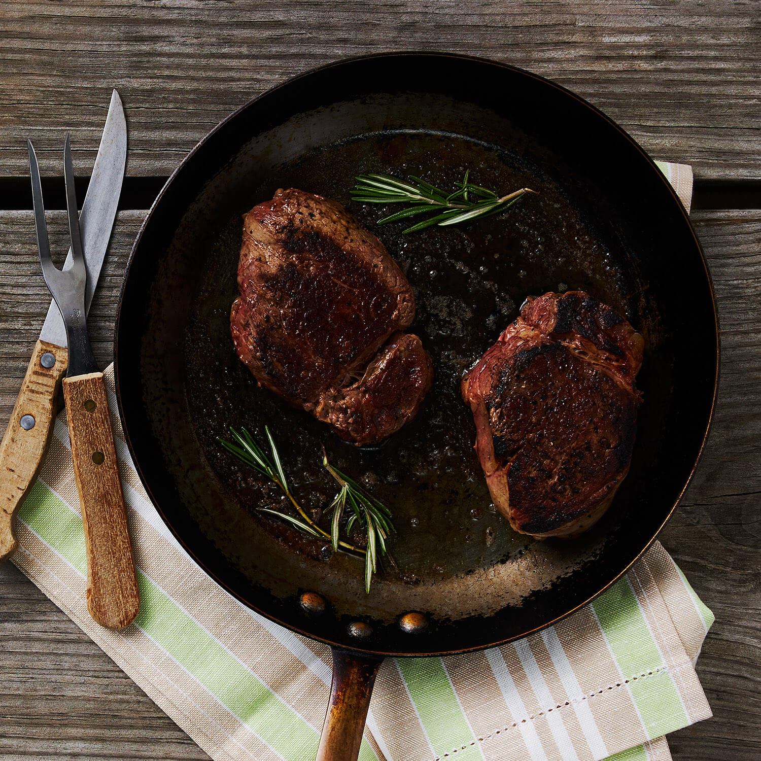 Two seared tenderloin steak fillets in a frying pan with rosemary on a wooden table.