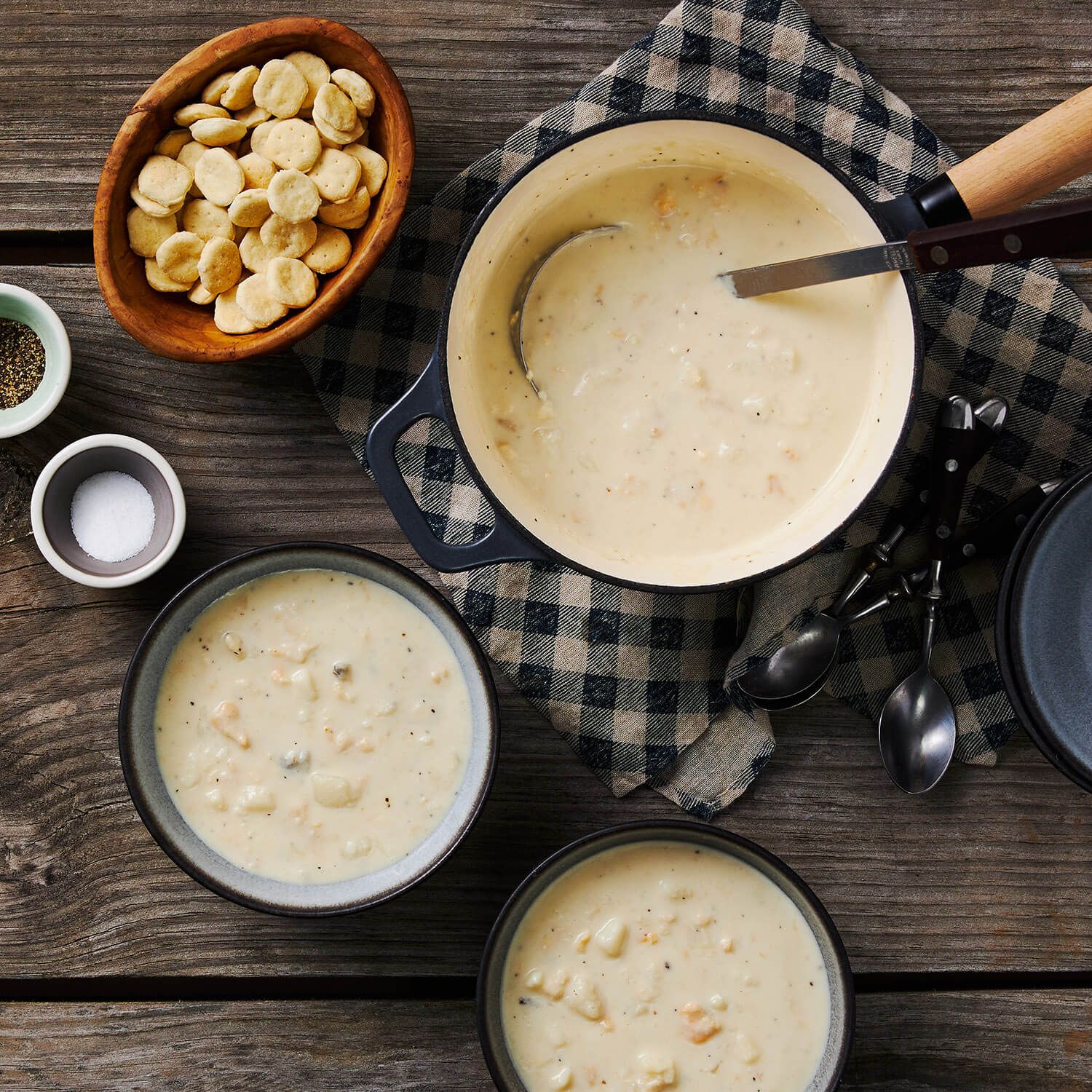 Pots and bowls of New England Clam Chowder on a wooden table with crackers and seasonings.