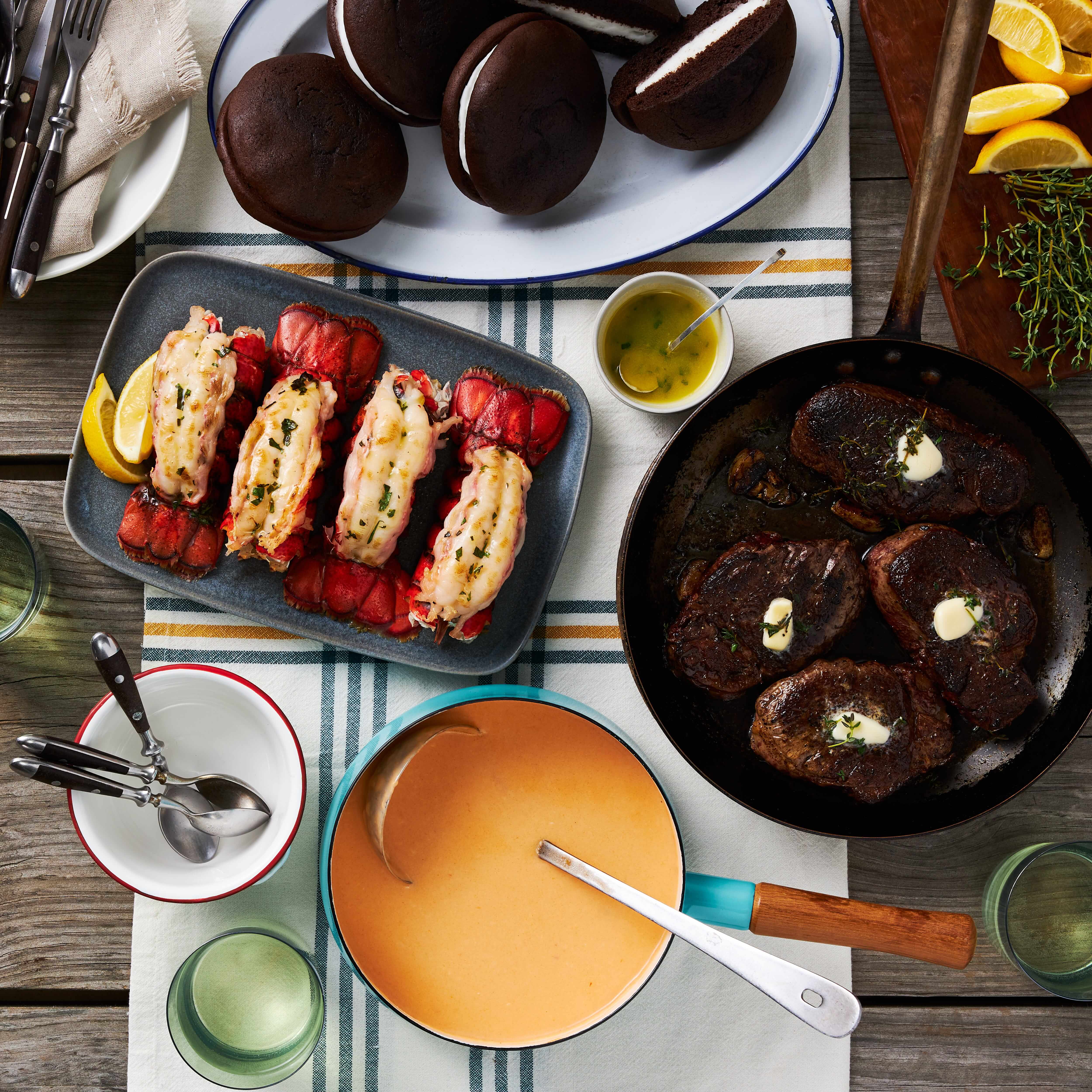 A variety of prepared dishes including Surf & Turf meal components such as lobster tails and steak, served on a table with sides like lobster bisque and whoopie pies.
