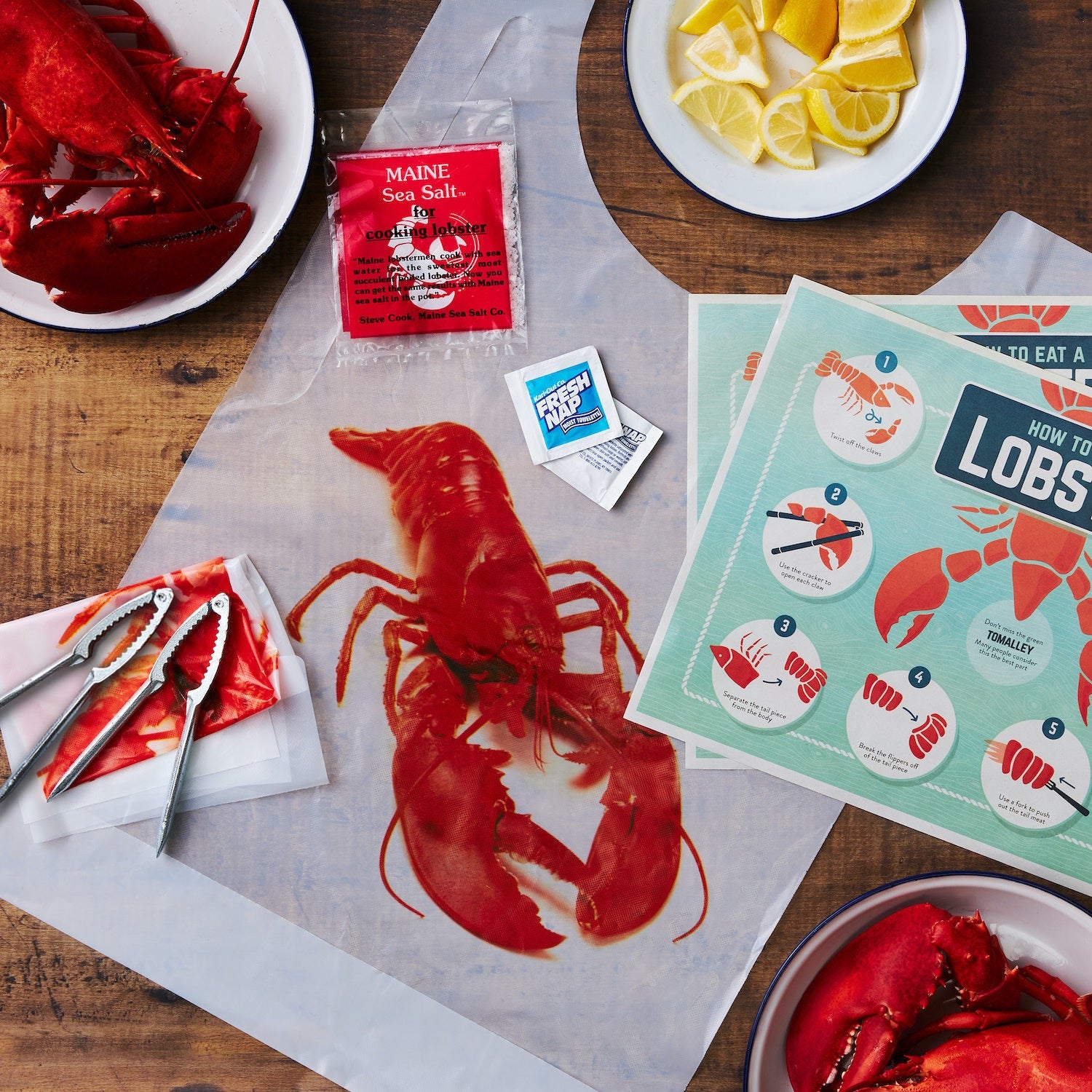 This photograph shows our Cousins Maine Lobster Toolkit laid out on a dining table beside some freshly steamed lobsters ready to be enjoyed! The kit includes How-to-eat a Lobster Placemats, Two Lobster Claw Cracker, a pair of Lobster Bibs, a Packet of Maine Sea Salt (to season pot when cooking), and two Wet Naps for cleaning up!