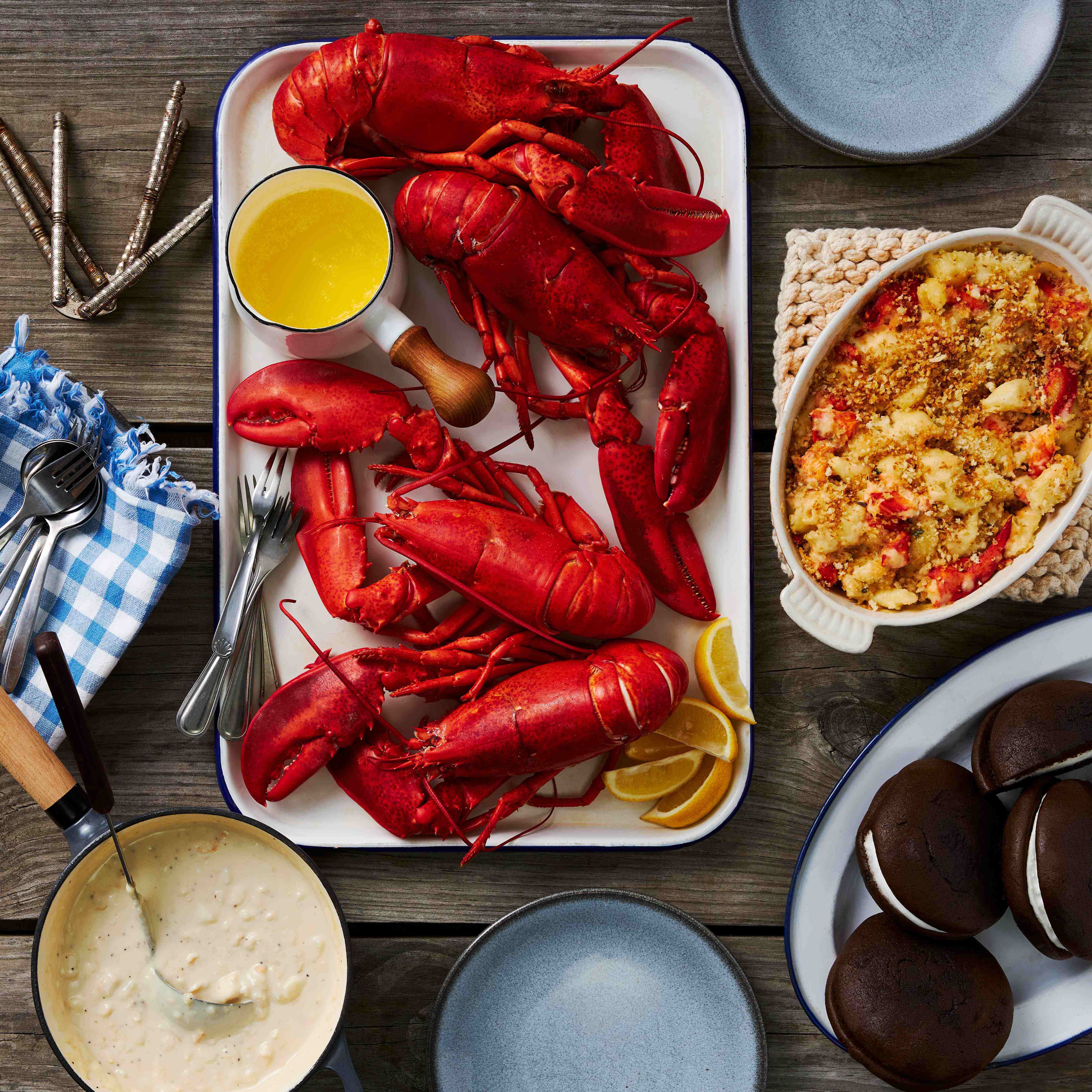 A variety of dishes including four red live Maine lobsters, a bowl of butter, a serving of Maine Lobster Mac and Cheese, a bowl of clam chowder, and four whoopie pies on a wooden table.
