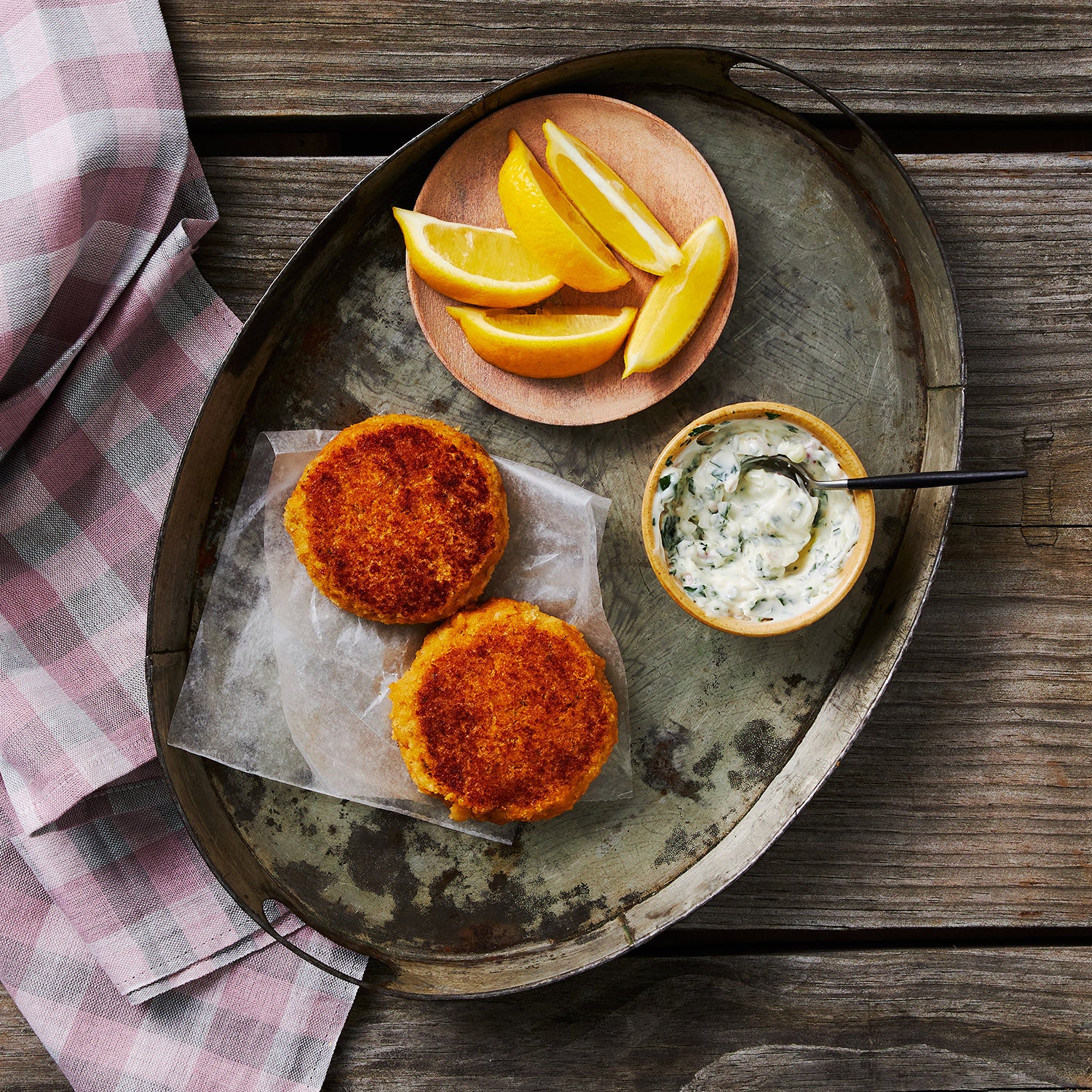 This photograph shows 2 crab cakes cooked to a perfect golden brown crust on a metal tray with lemon wedges and a herb aioli sauce to complete the meal