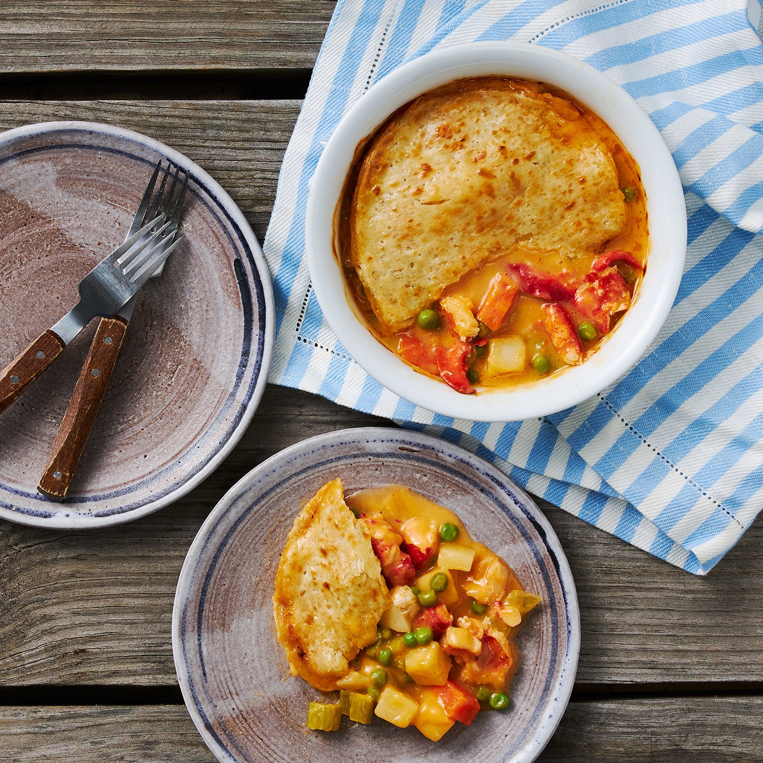 This Photo Shows a Maine Lobster Pot Pie fresh out of the oven with a slice taken out of it that has been plated on one of the two accompanying dishes.  Both the plate and the dish the Pot pie were prepared with are teeming with the filling of Maine Lobster, Potatoes, Celery, Carrots, Peas and the creamy base.