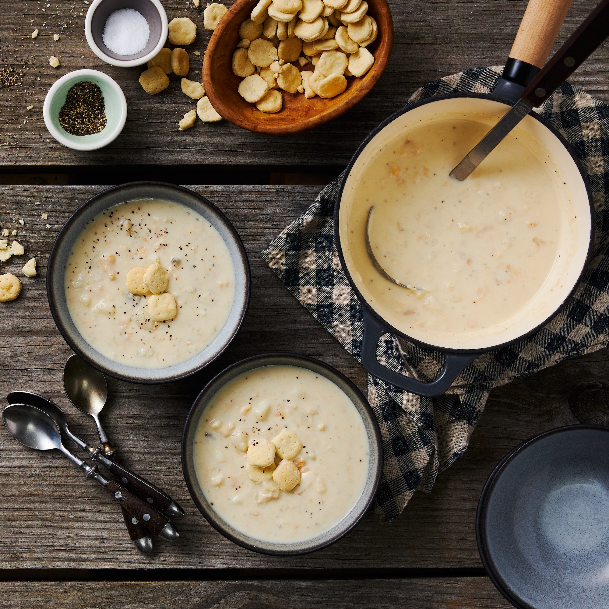 This image shows a large pot of New England Clam Chowder, accompanied by two full bowls seasoned with cracked pepper and topped with oyster crackers.