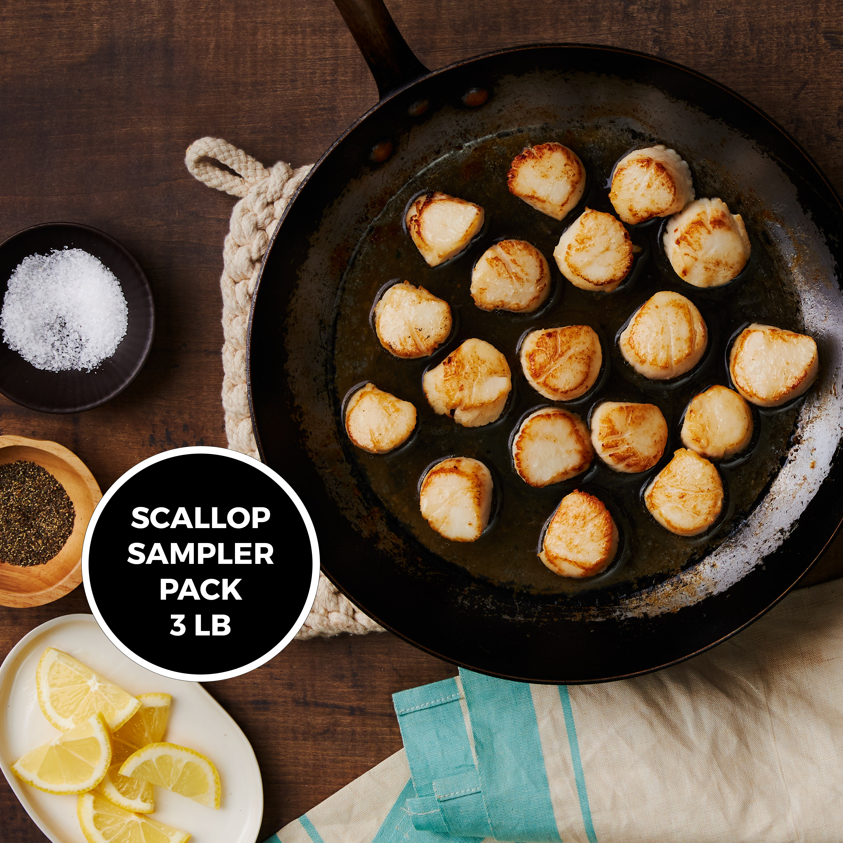 This Image shows 18 Maine Dayboat scallops freshly cooked in a cast iron pan, surrounded by a dishcloth, salt and pepper and lemon wedges.