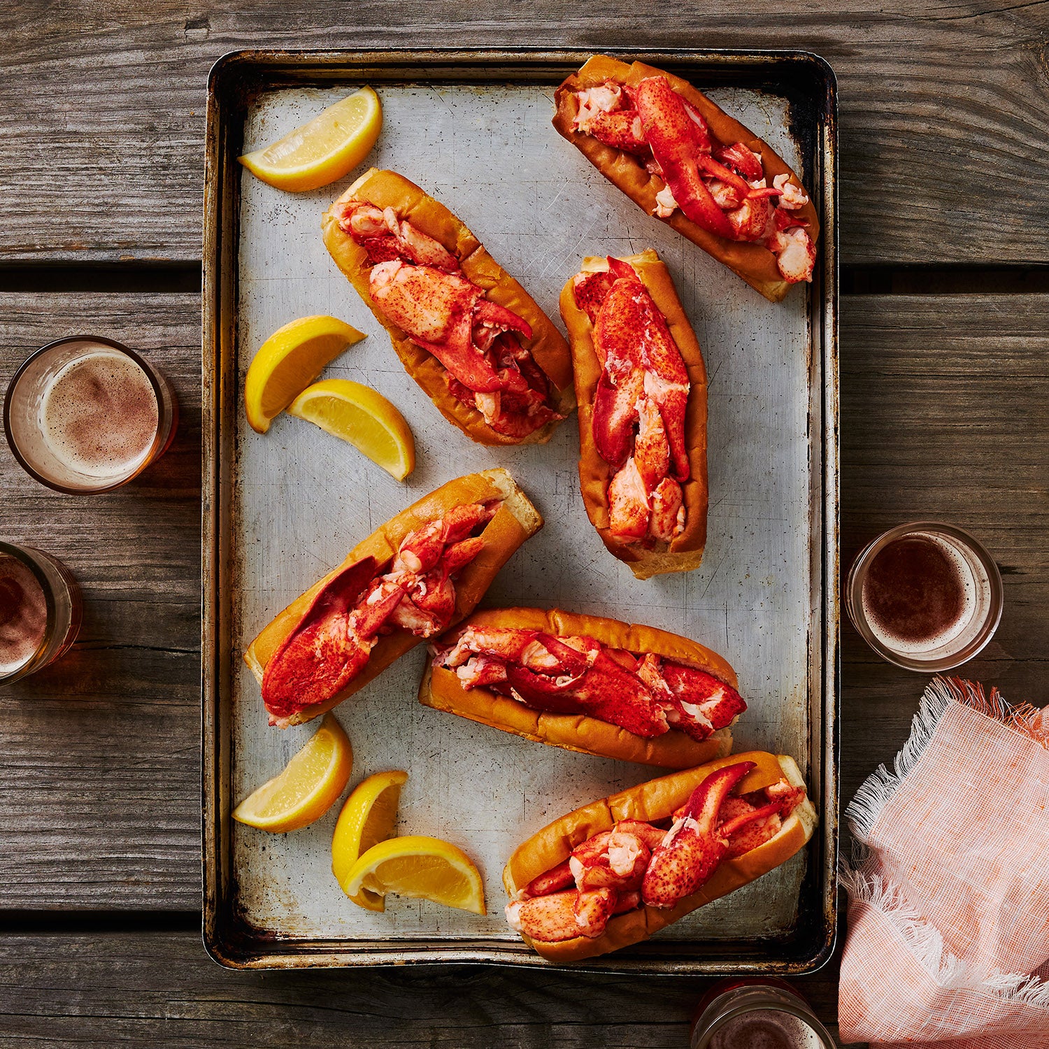 This image depicts 6 Lobster Rolls teeming with mouth watering Maine Lobster meat, upon a Baking sheet with wedges of Lemon to be squeezed overtop laid about the tray.