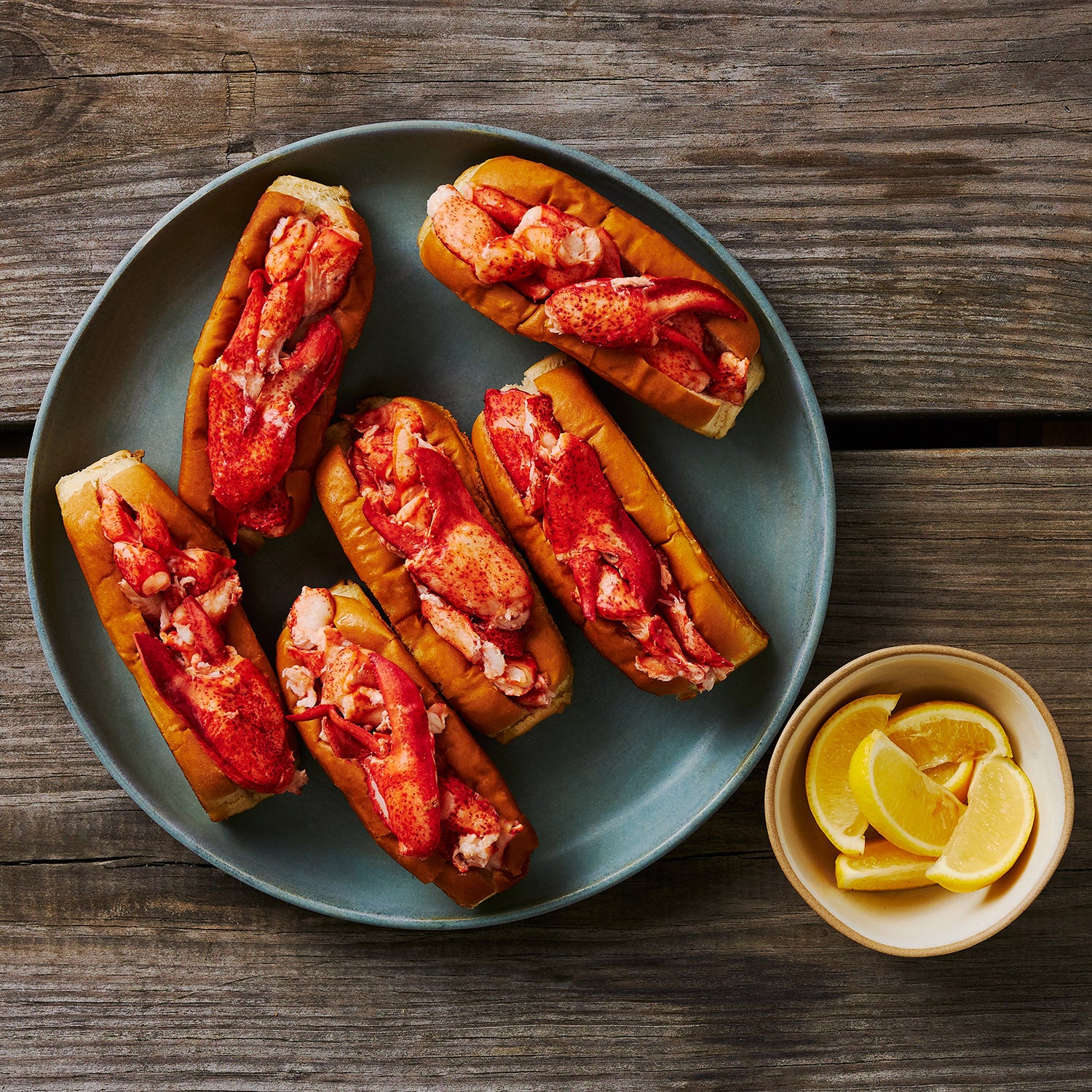 This Photograph shows 6 Maine Lobster Rolls plated and ready to be enjoyed with an accompanying bowl of fresh sliced lemon wedges.  